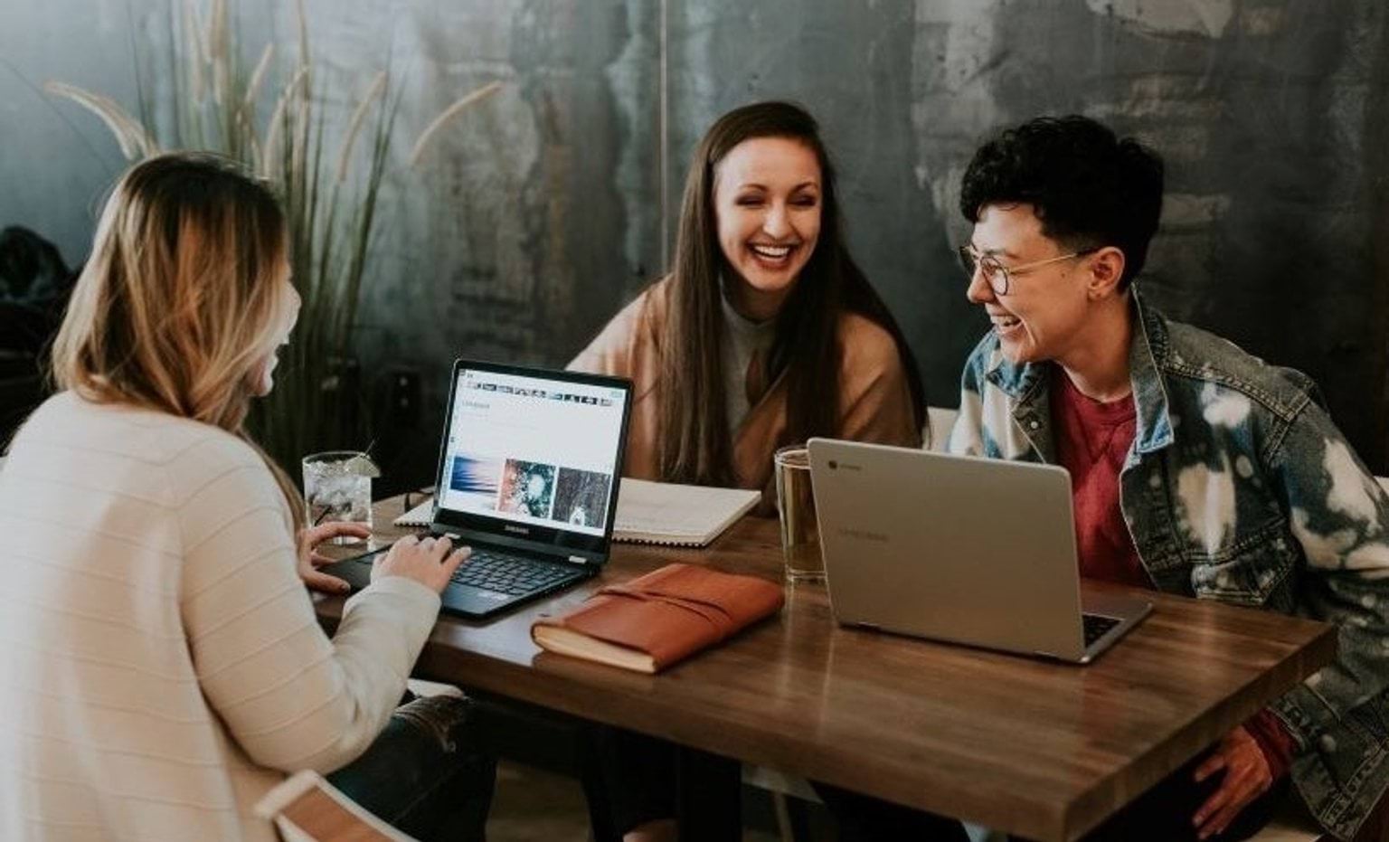 Three people are sitting at a table smiling and laughing. There are two laptops on the table.