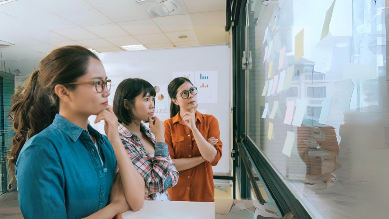 3 people looking at a board of post it notes. They each have one of their hands holding their chin in a thinking posture.