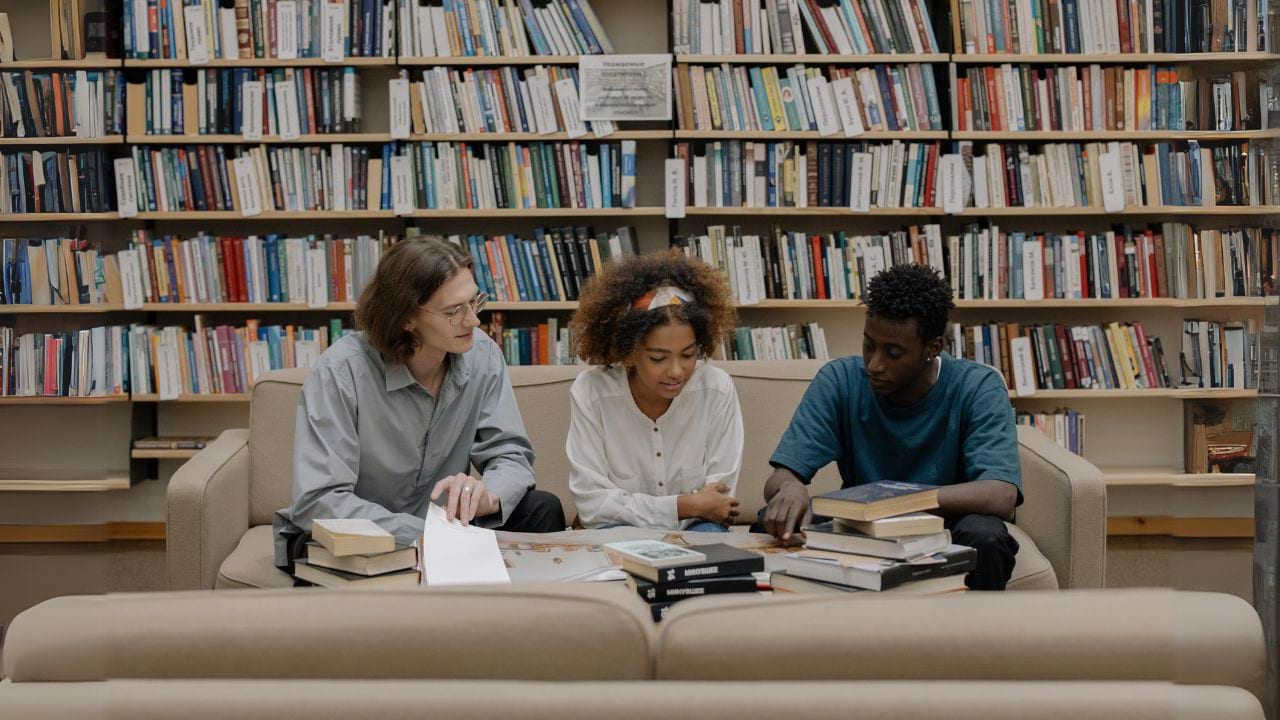 3 people sitting on a couch in front of a long and tall bookshelf that is full of books. The three people have a table in front of them, with multiple books stacked on top of it. They have one book open in front of the three of them and one person is pointing at the book, while the other two look at what is being pointed at.