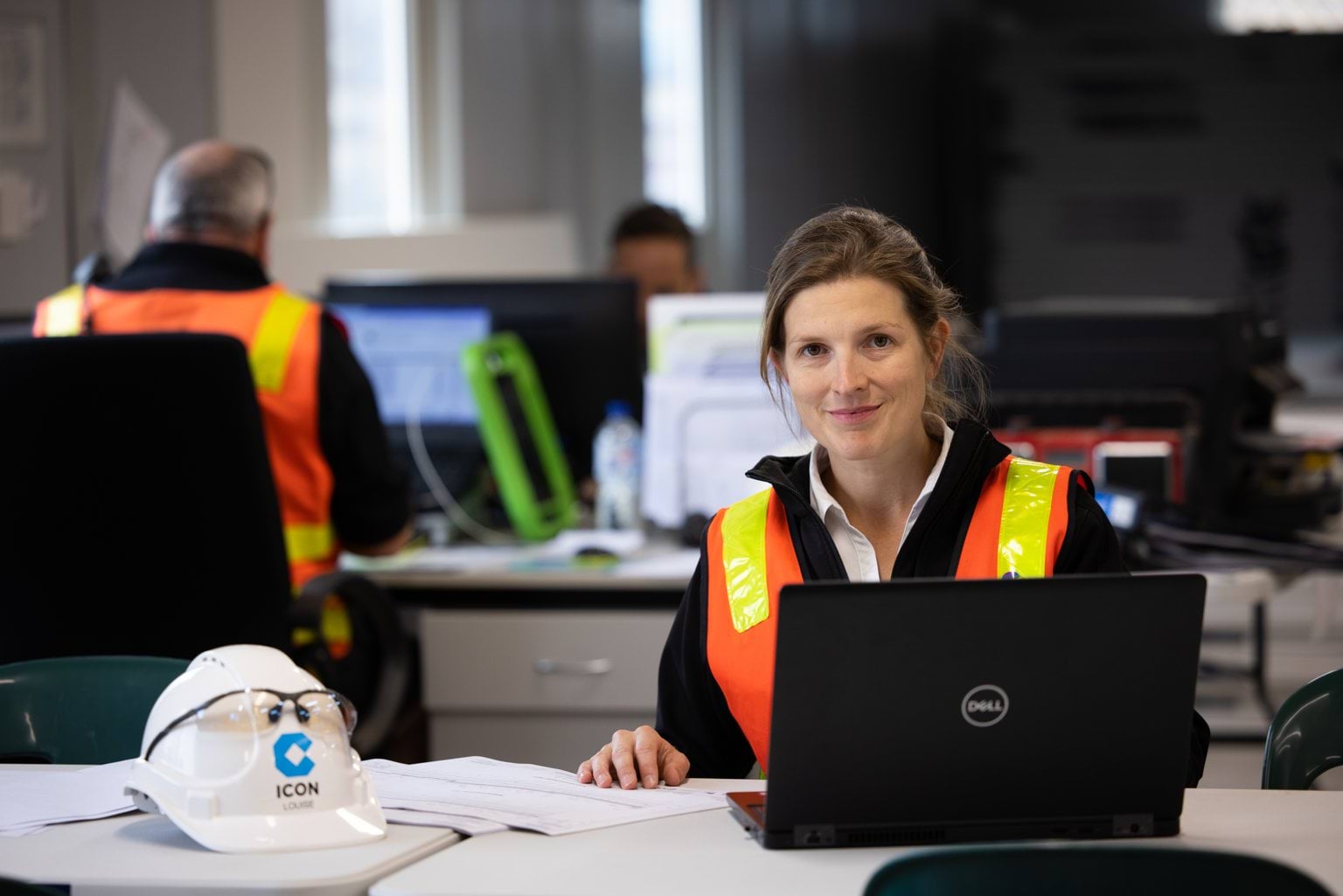 Person in a high-visibility vest working on a laptop at a construction office desk, with colleagues in the background